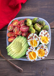 eggs, avocado, tomatoes, and brussel sprouts in a container on a wood piece.