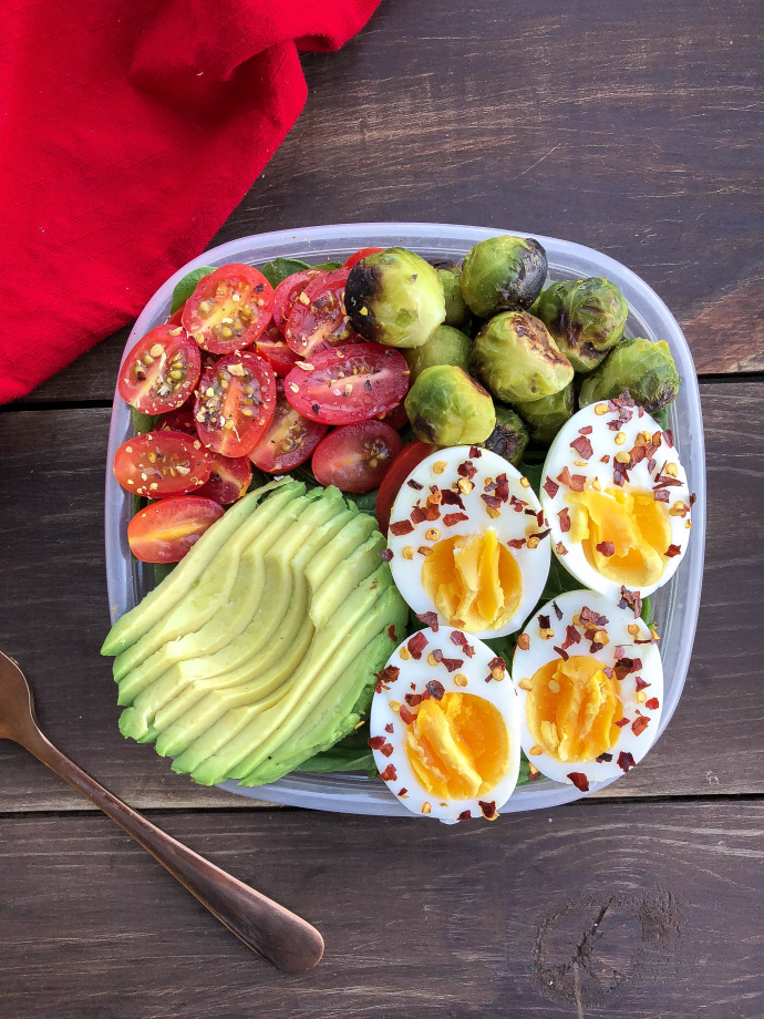 eggs, avocado, tomatoes, and brussel sprouts in a container on a wood piece. 