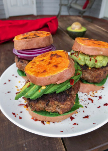 Grass fed burger in between sweet potato buns on a white plate with red pepper flakes.