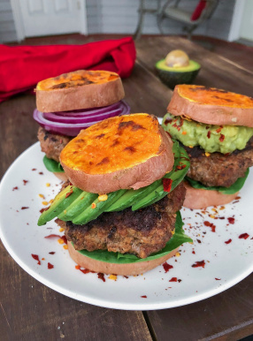 Grass fed burger in between sweet potato buns on a white plate with red pepper flakes.