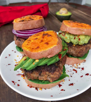 Grass fed burger in between sweet potato buns on a white plate with red pepper flakes.