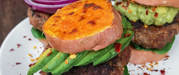 Grass fed burger in between sweet potato buns on a white plate with red pepper flakes.