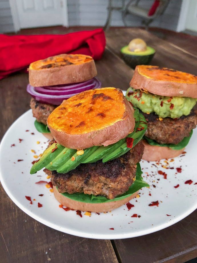 Grass fed burger in between sweet potato buns on a white plate with red pepper flakes. 