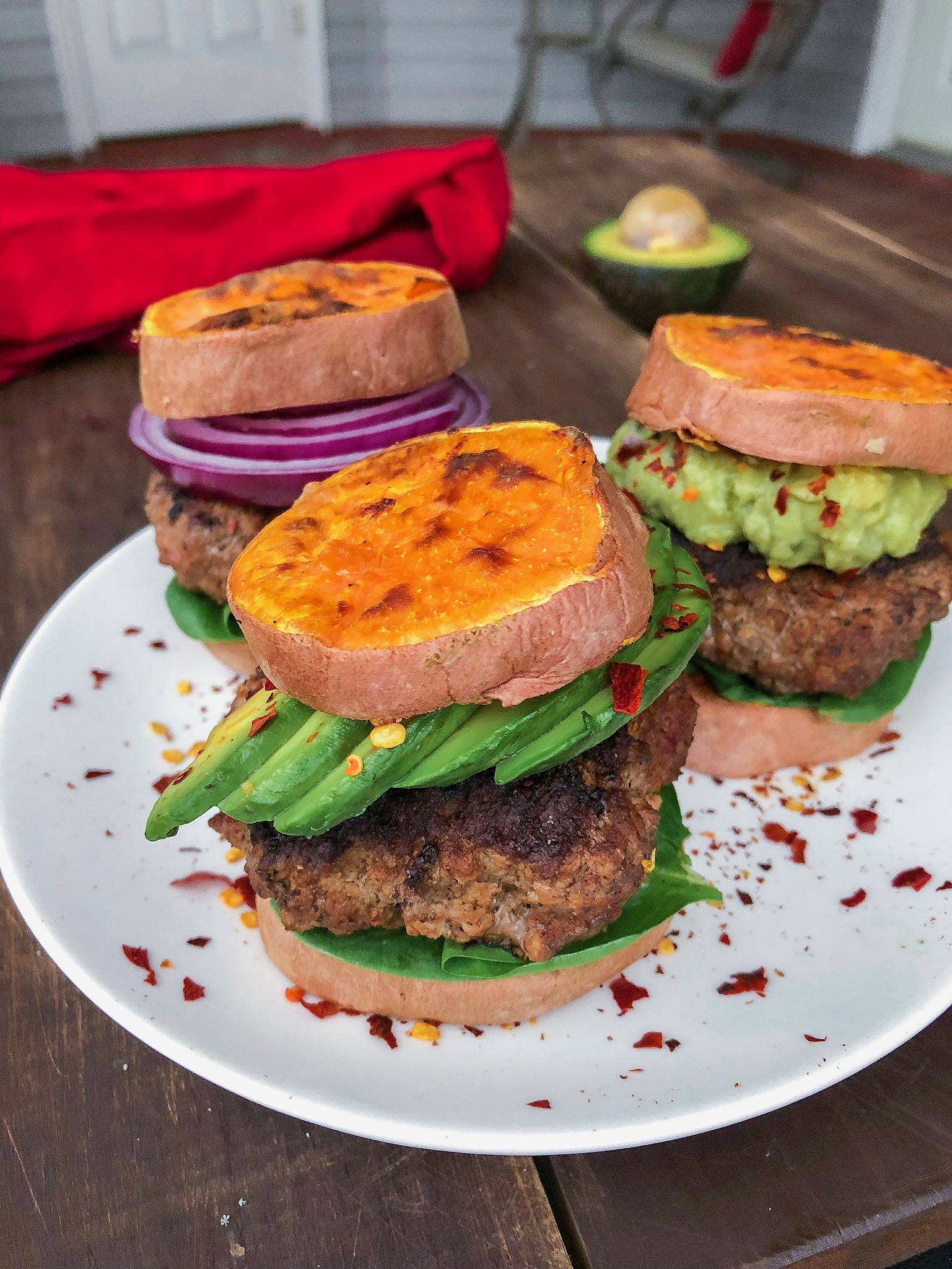 Grass fed burger in between sweet potato buns on a white plate with red pepper flakes.