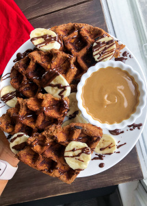 paleo sweet potato waffle on a white plate with cashew butter, bananas, and chocolate drizzled on top