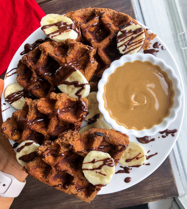 paleo sweet potato waffle on a white plate with cashew butter, bananas, and chocolate drizzled on top