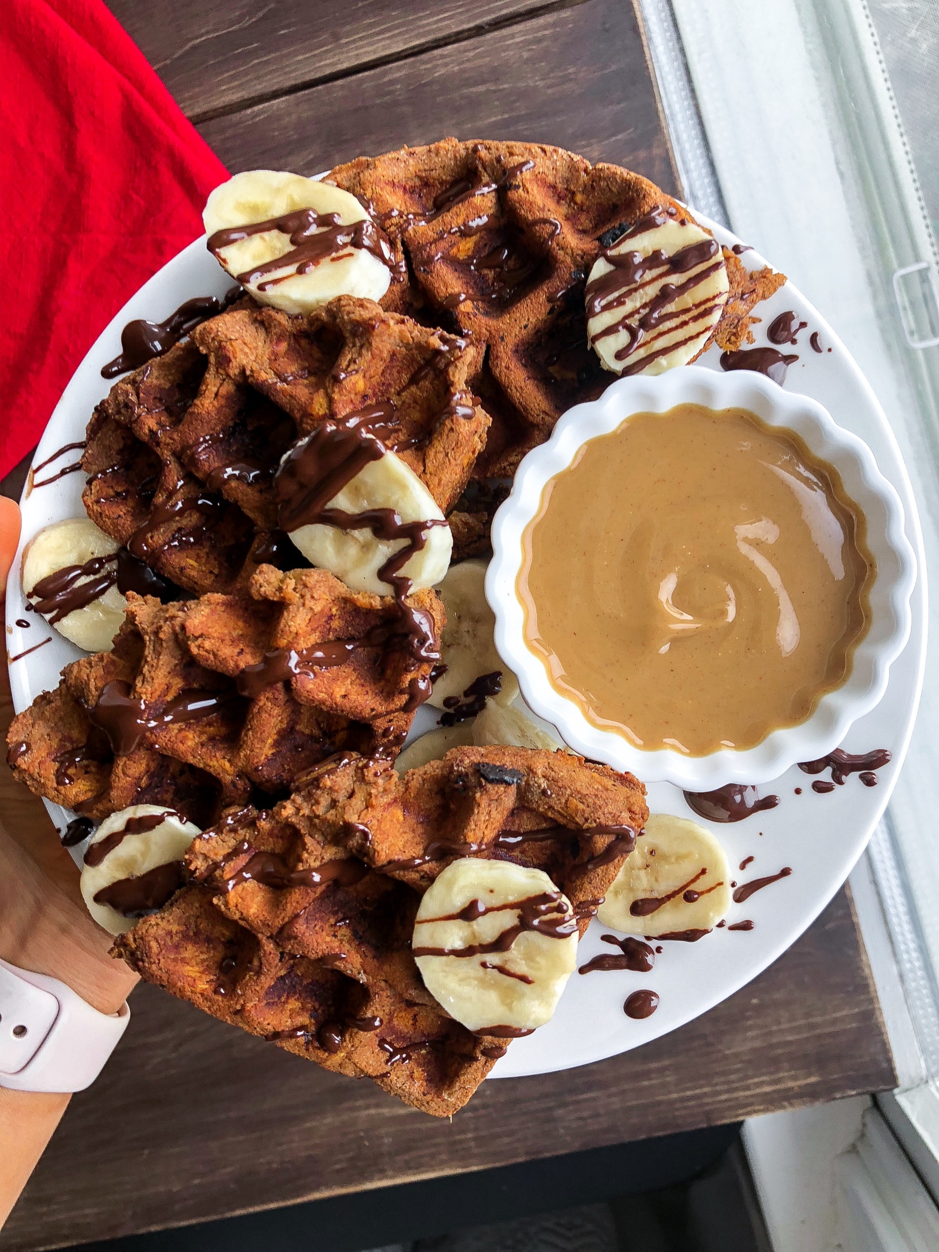paleo sweet potato waffle on a white plate with cashew butter, bananas, and chocolate drizzled on top