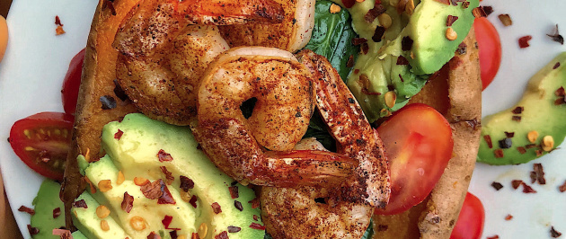 Shrimp, avocado, tomatoes, and spinach stuffed in a sweet potato on a white plate.