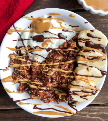 Chocolate oatmeal in a white bowl with banana, cashew butter, yogurt, and chocolate drizzled on top