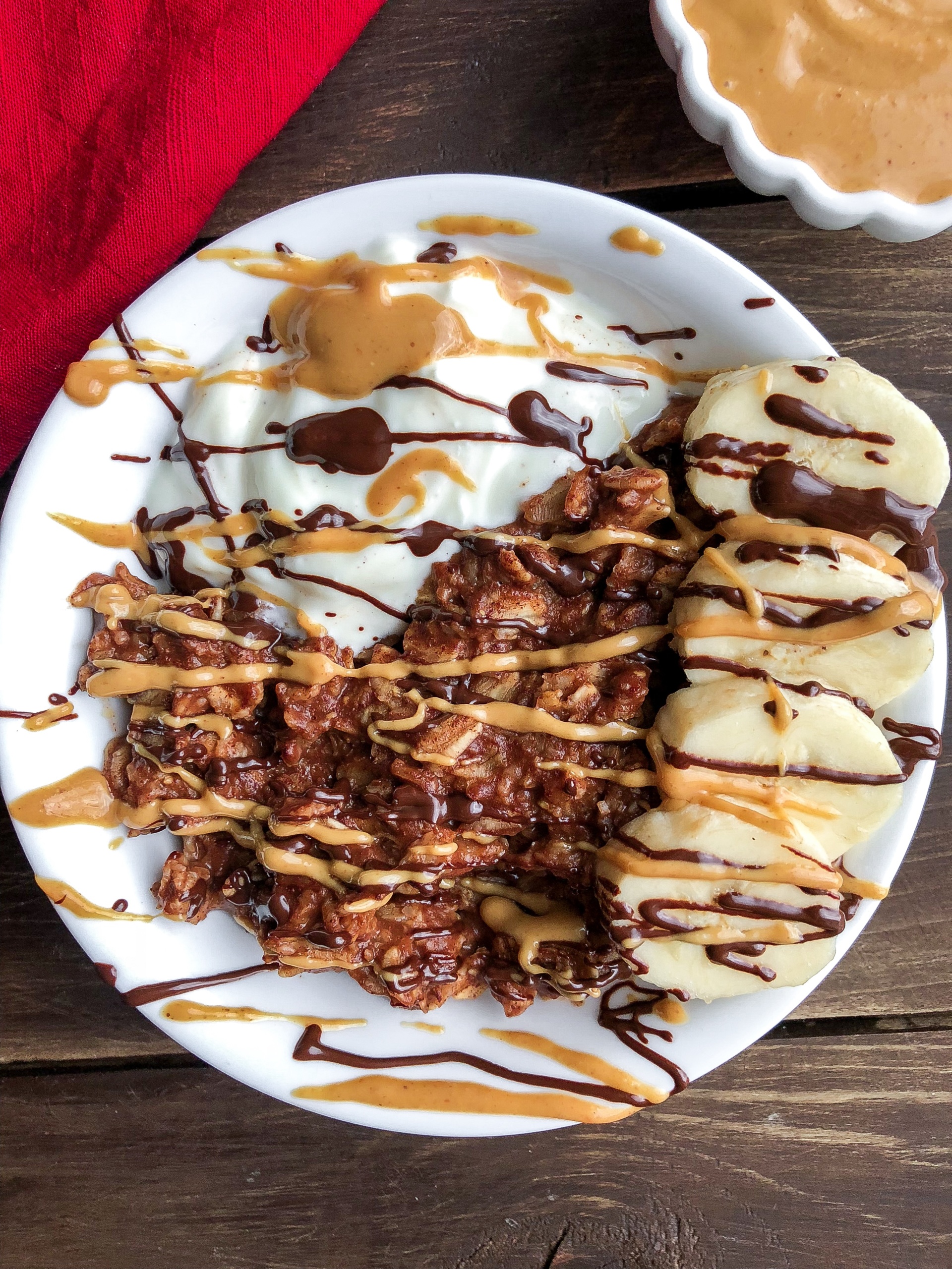 Chocolate oatmeal in a white bowl with banana, cashew butter, yogurt, and chocolate drizzled on top